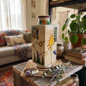 May include: A square ceramic vase with a cream-colored crackle glaze finish, featuring hand-painted motifs of a yellow sun, green triangular mountains, and small flowers. The vase has a flared neck and base with colorful geometric accents.