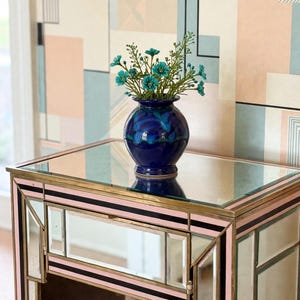 May include: A small, round, deep blue ceramic vase containing a bouquet of small teal flowers. The vase sits on a mirrored console table with gold-toned metal trim, pink accents, and black stripes, featuring a reflective glass top surface.