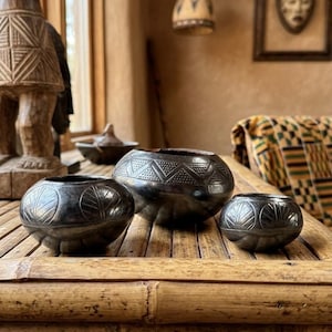 May include: Three rounded black ceramic bowls with polished surfaces and etched geometric patterns. The bowls vary in size, featuring circular leaf-like motifs on the smaller vessels and a zigzag band with dotted textures on the central, largest bowl.
