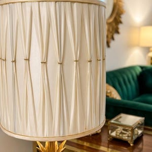 May include: A cylindrical lamp shade in an off-white cream fabric featuring vertical pleats secured with small gold-toned accents. The shade sits atop a gold-colored base shaped like a pineapple, with a smooth, structured fabric finish.