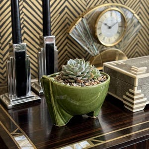May include: A small, glossy olive green ceramic planter with three short, angled feet, containing a light green succulent plant nestled in a bed of small, multi-colored pebbles. The planter has a smooth, curved bowl shape.