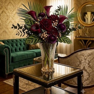 May include: A floral arrangement featuring deep burgundy calla lilies, dark red dahlias, and purple ranunculus flowers. The bouquet includes green palm fronds, eucalyptus leaves, and small metallic gold berries, all displayed in a ribbed glass vase.