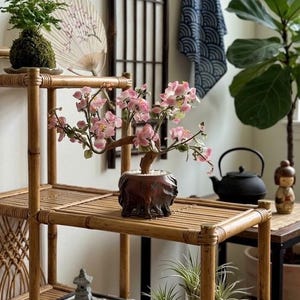 May include: A decorative bonsai tree sculpture with light pink blossoms and a brown trunk, set in a dark carved wooden pot. The sculpture sits on a bamboo shelf alongside a small zen garden tray with stones and a miniature stone pagoda statue.