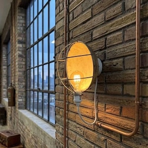 May include: A wall-mounted light fixture featuring a round, bowl-shaped shade with a protective metal wire cage. The lamp contains a warm-glowing Edison-style light bulb and is connected to a white power cord, mounted on a textured brick wall.