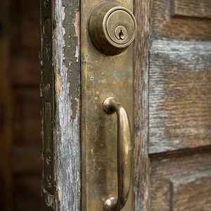 May include: A weathered brass door handle and deadbolt lock assembly mounted on a textured, dark brown wooden door. The metal hardware features a tarnished, aged patina with visible signs of wear, including a rectangular backplate and a curved grip.