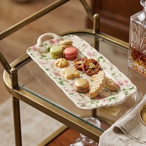 May include: A rectangular white ceramic serving tray with a floral pattern of pink flowers and green vines, featuring two twisted handles. The tray holds an assortment of colorful macarons, small cookies, and biscotti on a brass bar cart.