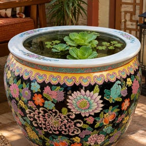 May include: A large, round ceramic fish bowl featuring a black background decorated with colorful floral patterns in pink, yellow, orange, and green. The wide, white rim has a delicate floral border, and the interior contains water with floating green aquatic plants.
