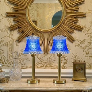May include: A pair of table lamps featuring brass-toned metal bases and vibrant blue glass shades with a textured, geometric diamond pattern. The shades have a scalloped bottom edge and are illuminated from within, casting a bright blue glow.