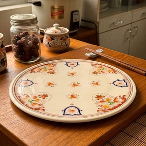 May include: A round ceramic plate with a cream-colored base, featuring a repeating pattern of orange and yellow floral motifs, accented by dark blue shield-shaped borders and intricate red filigree details along the outer rim of the plate.