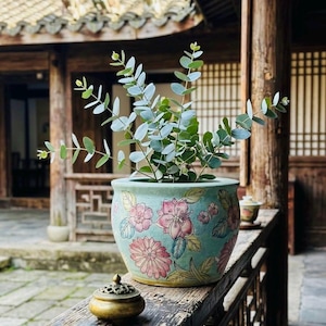 May include: A light blue ceramic planter featuring hand-painted pink and red floral motifs with green leaves, containing a green eucalyptus plant with rounded leaves. A small brass incense burner with a perforated lid sits nearby.