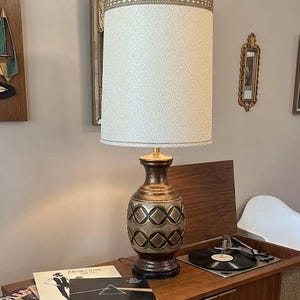 May include: A table lamp with a textured off-white cylindrical shade featuring a decorative gold trim at the top. The base is a bulbous ceramic vessel with a metallic bronze finish, accented by black and gold geometric diamond patterns.