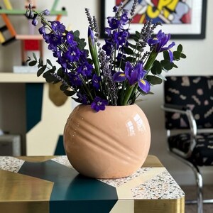 May include: A round, peach-colored ceramic vase with diagonal embossed ridges, filled with a vibrant bouquet of deep purple delphiniums, lavender sprigs, blue irises, and green eucalyptus leaves, creating a striking contrast of colors.