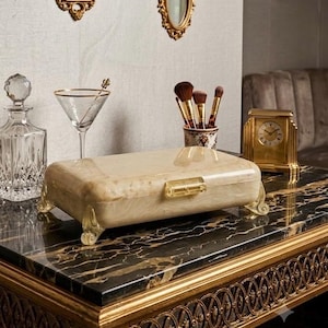 May include: A rectangular cream-colored box with a marbled finish, featuring a clear acrylic latch and four curved, translucent feet. The box has a smooth, polished surface and is designed with a hinged lid for storage.