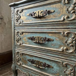 May include: A three-drawer chest featuring a distressed teal and gold finish. The wooden furniture piece displays ornate carved molding along the drawer fronts and edges, complemented by curved brass-toned metal handles on each drawer.