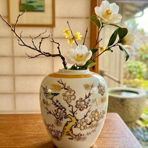 May include: A white ceramic vase with a gold rim and a hand-painted design featuring a yellow and brown flowering tree branch with a small green bird. The vase contains a bare twig, small yellow blossoms, and white camellia flowers.