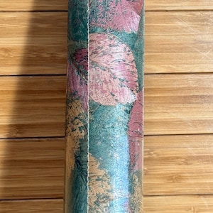 May include: A roll of wallpaper featuring a repeating pattern of stylized leaves in shades of teal, pink, and gold. The leaves have a textured appearance, and the background is a darker teal color. The roll is cylindrical.
