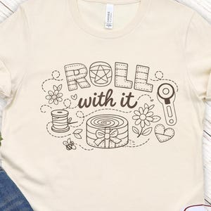 Puede incluir: Camiseta color crema con la frase "Roll with it" en una fuente caprichosa y cosida, acompañada de ilustraciones temáticas de costura. La camiseta se combina con pantalones cortos de mezclilla y zapatillas blancas, sobre un fondo de madera clara.