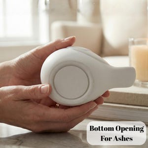 May include: A white ceramic urn with a rounded body and a spout, held in two hands. The bottom of the urn has a large circular opening. Text on the image reads "Bottom Opening For Ashes".
