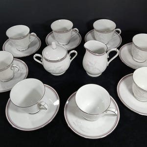 May include: A white porcelain tea set featuring six teacups with matching saucers, a lidded sugar bowl with two handles, and a creamer pitcher. Each piece is decorated with a subtle, light gray leaf pattern and finished with a thin silver rim.