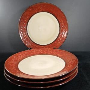 May include: A set of four round ceramic dinner plates featuring a cream-colored center and a wide, dark red rim. The rim is decorated with a raised, embossed scrollwork pattern and a dark brown edge, creating a distinct contrast between the colors.