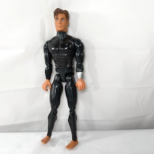 May include: A black action figure with a muscular torso, wearing a black suit with a high collar. The figure has brown hair, tan skin, and a silver wristband. The figure is standing on a white background.