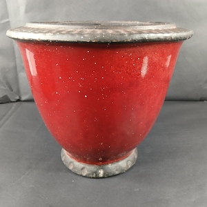 May include: A red ceramic bowl with a speckled finish and a decorative, dark metal rim. The bowl has a wide, rounded shape and sits on a matching metal base. This vintage bowl is suitable for kitchen decor.