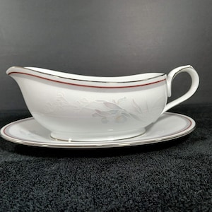 May include: A white porcelain gravy boat with an attached saucer, featuring a thin red rim and a subtle, light-colored floral pattern embossed on the side. The piece has a curved handle and a smooth, glossy finish with a silver-toned trim.