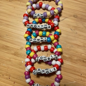 May include: A collection of vibrant beaded bracelets. Each bracelet displays a unique colour palette and incorporates letter beads spelling out names such as "Jax", "Ragatha", "Pomni", "Zooble", "Gangle", and "Kinger". The beads vary in size and shape.