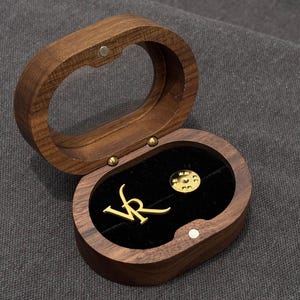 Könnte beinhalten: Eine offene, ovale Schmuckschatulle aus Holz mit einer goldfarbenen Anstecknadel und den Buchstaben "VR" im Inneren. Die Schatulle ist aus dunklem Holz gefertigt und hat einen Magnetverschluss. Der Innenraum ist mit schwarzem Samt ausgekleidet.