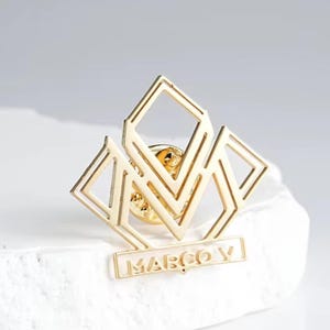 Puede incluir: Un pin dorado con un diseño geométrico con formas de diamante y el texto "MARCO V". Este elegante pin es un gran accesorio, ideal para añadir un toque de estilo a la ropa o accesorios.