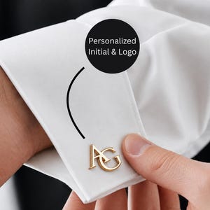 May include: A gold-toned metal cufflink featuring the letters AG attached to the cuff of a crisp white dress shirt. A black circular graphic overlay with the white text Personalized Initial & Logo points to the gold cufflink.