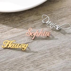 Könnte beinhalten: Drei personalisierte Namensbroschen in Gold, Roségold und Silber. Die Namen "Harvey", "Sophia" und "Daniel" sind in einer Schreibschrift dargestellt. Die Broschen sind auf einer Holzoberfläche angeordnet, mit einem weißen Teller im Hintergrund.