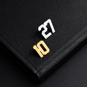 May include: Two metal number pins, one silver with the number 27 and one gold with the number 10, are displayed on a black leather surface. The numbers are in a bold, sans-serif font, creating a modern aesthetic.