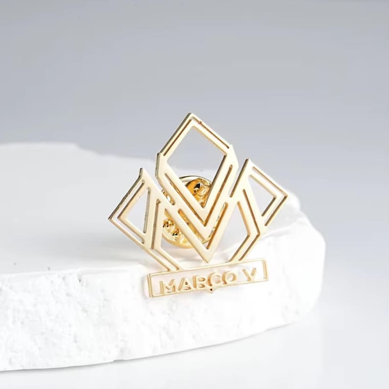 K&ouml;nnte beinhalten: Eine goldfarbene Anstecknadel mit einem geometrischen Design mit Diamanten und dem Text "MARCO V". Diese Anstecknadel ist ein stilvolles Accessoire, perfekt, um Kleidung oder Accessoires einen Hauch von Flair zu verleihen.