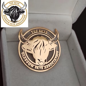 Puede incluir: Un pin metálico dorado con la cabeza de una vaca de las Tierras Altas dentro de un marco circular. El texto "THE SKYE" está arqueado en la parte superior y "GLASGOW SKYE ASSOCIATION" en la inferior. El pin se muestra en una caja blanca.