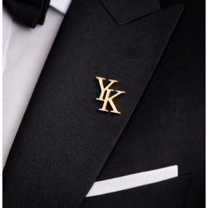 May include: A gold-colored lapel pin with the intertwined letters "YK" is affixed to a black suit jacket. The jacket has a white pocket square and is paired with a white dress shirt and a black bow tie.