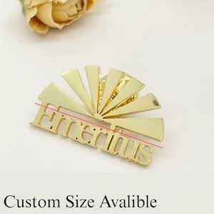 K&ouml;nnte beinhalten: Eine goldfarbene Anstecknadel mit dem Wort "Emeritus" in einer stilisierten Schriftart. Die Anstecknadel hat ein Sonnenstrahl-Design hinter dem Text. Der Text "Custom Size Avalible" steht unter der Anstecknadel.
