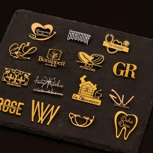 K&ouml;nnte beinhalten: Eine Sammlung goldfarbener Metallnadeln und Broschen mit verschiedenen Designs und Texten. Die Nadeln umfassen ein Herz, ein stilisiertes Wort, ein medizinisches Symbol, ein Logo und die Buchstaben "GR" und "WW". Sie sind auf einer dunklen, strukturierten Oberfl&auml;che angeordnet.