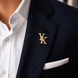 Puede incluir: Un pin de solapa de metal dorado con las letras YK en una fuente con serifa, sujeto a la solapa de una americana azul marino oscuro sobre una camisa blanca de cuello rígido con un pañuelo de bolsillo blanco en el pecho.