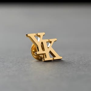 Może przedstawiać: Złota przypinka z przeplatającymi się literami "YK". Przypinka ma błyszczące wykończenie i jest umieszczona na szarym tle. Przypinka to stylowy dodatek.