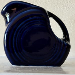May include: A dark blue ceramic pitcher with a unique, curved design. The pitcher has a handle and a spout, and the surface features a swirling pattern. The glossy finish reflects light, highlighting the deep color and shape.