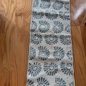 Puede incluir: Camino de mesa rectangular con cuentas con un patrón repetido de diseños de conchas marinas azules y blancas. El camino de mesa mide aproximadamente 30 cm de ancho y 91 cm de largo, con un fondo neutro y una superficie texturizada.