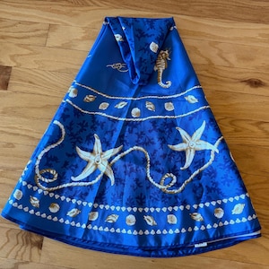 Puede incluir: Una tela decorativa azul con temática náutica. La tela presenta estrellas de mar, conchas marinas e hipocampos en blanco y dorado sobre un fondo azul. La tela está doblada sobre una superficie de madera.