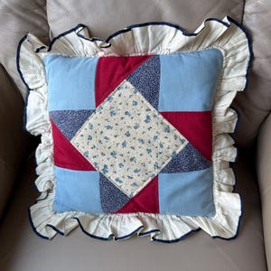 Könnte beinhalten: Ein quadratisches Zierkissen mit einem zentralen Sternmuster in Creme mit kleinem blauem Blumenmuster, umgeben von einfarbigen roten und hellblauen Stoffabschnitten. Das Kissen ist mit einem cremefarbenen Rüschenrand mit dunkelblauem Saum versehen.