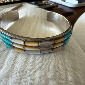 May include: A silver cuff bracelet featuring three horizontal rows of inlaid stones. The top and bottom rows consist of bright turquoise segments, while the middle row alternates between white and yellow rectangular stone inlays.