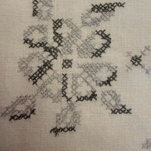 May include: Close-up of a white fabric with a cross-stitch floral design. The embroidery features gray and black thread, creating a detailed pattern. The fabric has a textured appearance.
