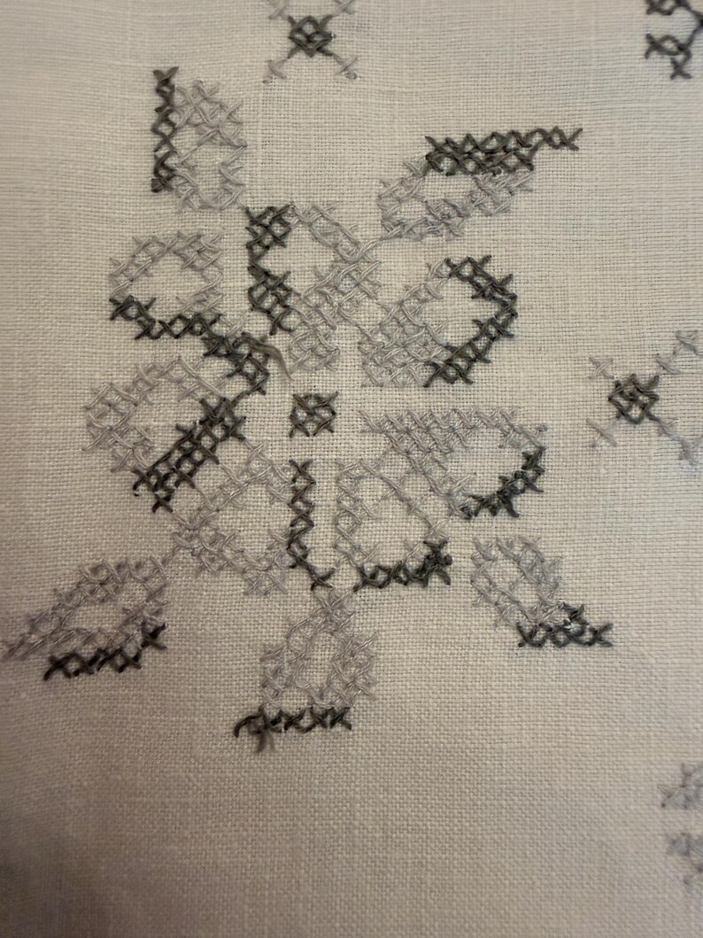 May include: Close-up of a white fabric with a cross-stitch floral design. The embroidery features gray and black thread, creating a detailed pattern. The fabric has a textured appearance.