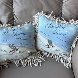 May include: Two decorative throw pillows with a beach theme. Each pillow features a light blue sky with the words "Paradise Found" in script. The pillows have a beach scene with a chair and seashells. The pillows are trimmed with a cream-colored rope.