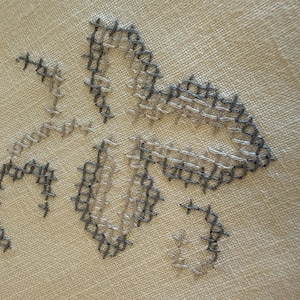 May include: Close-up of a cross-stitch floral design in shades of gray and beige. The embroidery features a stylized flower with detailed stitching on a neutral fabric background. The handmade craft showcases a delicate, textured pattern.