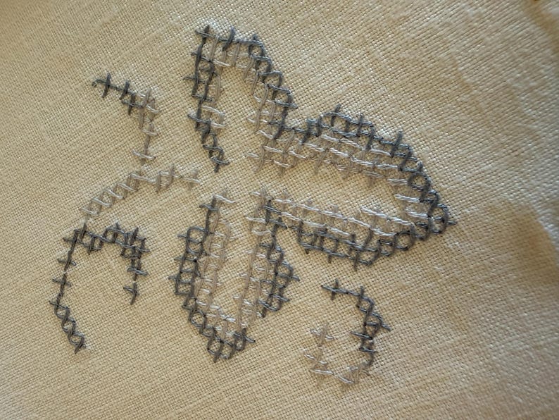 May include: Close-up of a cross-stitch floral design in shades of gray and beige. The embroidery features a stylized flower with detailed stitching on a neutral fabric background. The handmade craft showcases a delicate, textured pattern.